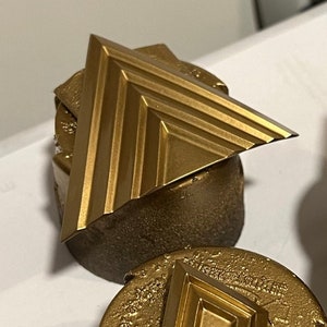 Puede incluir: Dos piezas decorativas de metal de color dorado. La primera pieza es un tri&aacute;ngulo con una superficie texturizada. La segunda pieza es un diamante con una superficie texturizada.