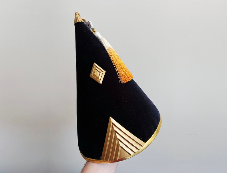 Puede incluir: Un sombrero de terciopelo negro en forma de cono con ribetes dorados y un borla dorada. El sombrero tiene un dise&ntilde;o geom&eacute;trico en la parte delantera.