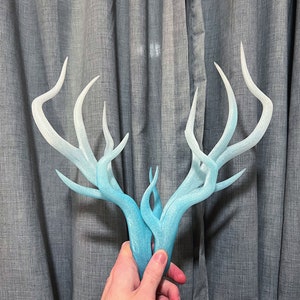 Può includere: Un paio di corna di cervo bianche e blu con un effetto sfumato. Le corna sono fatte di un materiale liscio e lucido e sono tenute nella mano di una persona.
