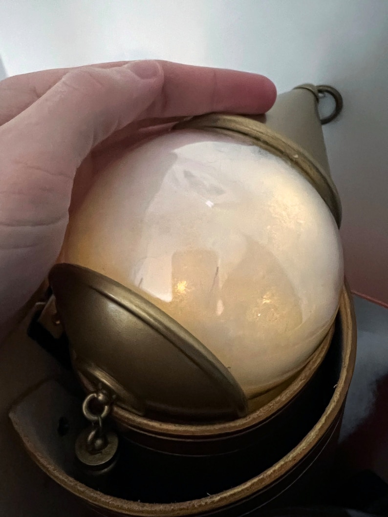 May include: A gold-colored, round, glass orb with a gold-colored metal lid and a chain attached. The orb is sitting on a brown leather base.