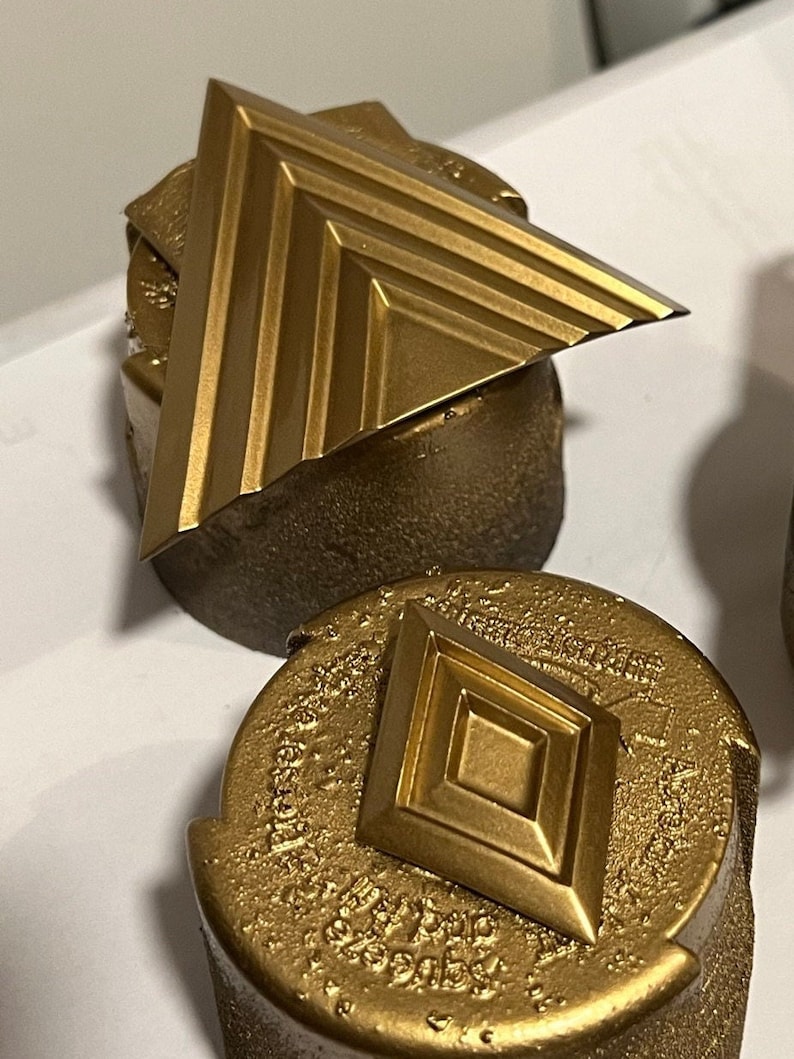 Puede incluir: Dos piezas decorativas de metal de color dorado. La primera pieza es un tri&aacute;ngulo con una superficie texturizada. La segunda pieza es un diamante con una superficie texturizada.