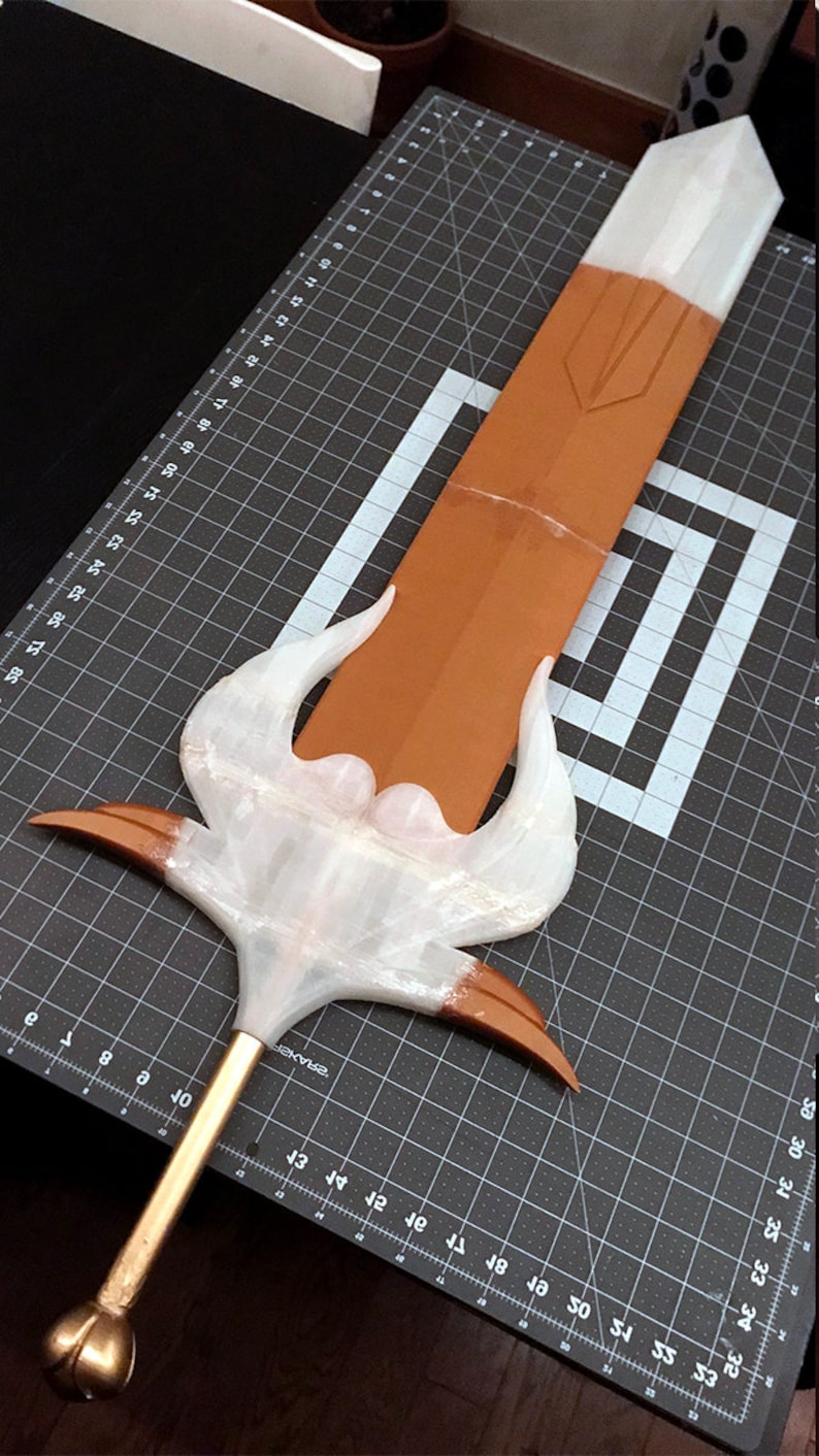Puede incluir: Una espada de fantas&iacute;a hecha a mano con una hoja marr&oacute;n y una empu&ntilde;adura blanca y dorada. La espada presenta un dise&ntilde;o estilizado con elementos curvos y un pomo redondeado. La hoja mide aproximadamente 61 cm de largo.