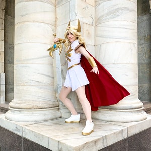 Puede incluir: Una persona con un disfraz de cosplay de She-Ra, sosteniendo una espada. El disfraz incluye un vestido blanco, una capa roja, detalles dorados y una corona dorada. La persona est&aacute; de pie frente a una columna de m&aacute;rmol.