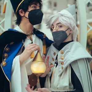 May include: Two people wearing elaborate costumes and face masks. One person is wearing a blue and gold robe with a tall black hat. The other person is wearing a white robe with a white hat and a black face mask. They are holding a small, glowing, gold orb.