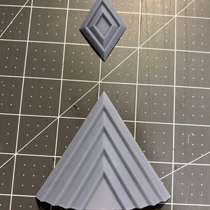 Puede incluir: Una forma geom&eacute;trica gris impresa en 3D que se asemeja a una pir&aacute;mide con una serie de crestas en cada lado. Tambi&eacute;n se muestra un objeto m&aacute;s peque&ntilde;o, gris, impreso en 3D, con forma de diamante con un centro cuadrado elevado.