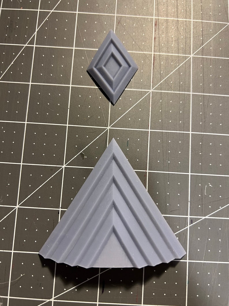 Puede incluir: Una forma geom&eacute;trica gris impresa en 3D que se asemeja a una pir&aacute;mide con una serie de crestas en cada lado. Tambi&eacute;n se muestra un objeto m&aacute;s peque&ntilde;o, gris, impreso en 3D, con forma de diamante con un centro cuadrado elevado.