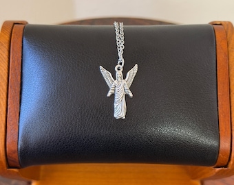 May include: A silver-colored angel pendant necklace. The pendant features an angel with outstretched wings and flowing robes. The necklace is displayed on a black leather surface with a wooden frame.