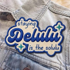May include: Embroidered patch with the text "staying Delulu is the solulu" in blue and white. The patch is in a cloud-like shape and is sewn onto a light blue denim jacket.