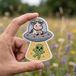 May include: Embroidered patch featuring a cow in a UFO, with a green alien below. The cow is brown and white, wearing a bell. The UFO is gray with orange accents. The alien is green with a yellow background.