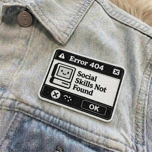 May include: A black and white patch with the text "Error 404 Social Skills Not Found" and a smiling computer icon. The patch is sewn onto a light blue denim jacket.