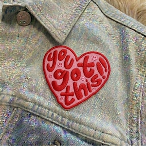 May include: Close-up of a light blue denim jacket with a shimmering, iridescent finish. A pink heart-shaped patch with the words "You got this!" in red embroidery is attached to the jacket. The patch includes small smiley faces.