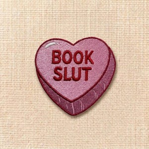 May include: Embroidered heart-shaped patch in shades of pink and red. The patch features the words "BOOK SLUT" in a bold, stitched font. The patch is likely for clothing or accessories.