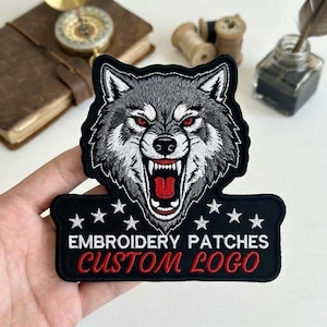 May include: Embroidered patch featuring a snarling wolf with red eyes and open mouth. The patch is gray, black, and white, with the text "EMBROIDERY PATCHES CUSTOM LOGO" in white and red.