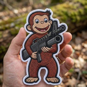 May include: Embroidered patch featuring a cartoon monkey holding a rifle. The monkey is brown with a tan face and a white border. The rifle is black and gray. The patch is held in a hand against a blurred green background.