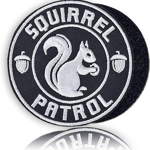 May include: Round black patch with white embroidered text and a squirrel graphic. The text reads "SQUIRREL PATROL" around the edge, with a squirrel in the center holding a nut. Two acorn graphics are on either side of the squirrel.