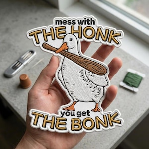 May include: Embroidered patch featuring a white duck holding a wooden paddle, with the text "mess with THE HONK you get THE BONK" in gold lettering with a white border. The patch is held in a hand.