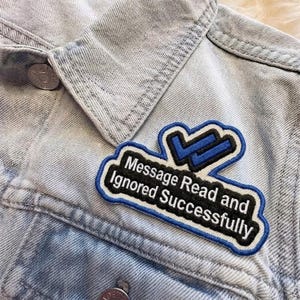 May include: A denim jacket with a patch that reads "Message Read and Ignored Successfully." The patch is black with white text and a blue checkmark design. The jacket is light blue.