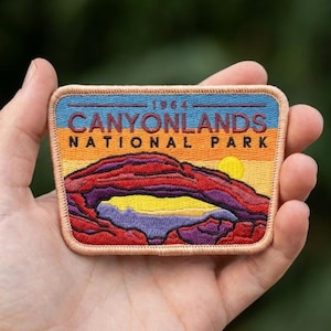 May include: A rectangular patch featuring the text "CANYONLANDS NATIONAL PARK" above a landscape illustration. The patch has a tan border and depicts a sunset scene with a red rock formation, a yellow sun, and purple and orange hues.