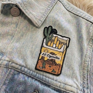 May include: A light blue denim jacket with a decorative patch. The patch features a desert scene with a pack of cigarettes, a cactus, and the text "20 Cigarettes".