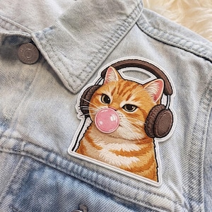 May include: Embroidered patch featuring a ginger tabby cat wearing brown headphones and blowing a pink bubble. The patch is on a light blue denim jacket. The cat has a serious expression.