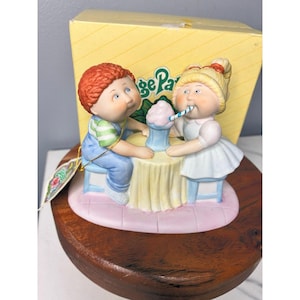 May include: A ceramic figurine depicting two children sharing a milkshake. The boy has red hair and blue overalls, the girl has blonde hair and a white dress. The figurine sits on a wooden base, with a yellow box in the background.