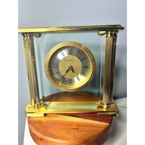 Relógio de mesa Howard Miller em latão e vidro com numerais romanos e mecanismo de quartzo.