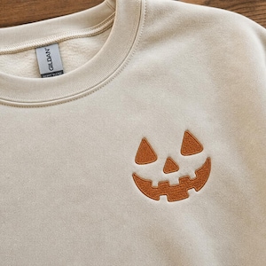 May include: A sand-colored crewneck sweatshirt featuring a small, embroidered orange jack-o'-lantern face on the left chest. The garment has a ribbed collar and a Gildan brand label visible on the inside of the neckline, showcasing a simple design.