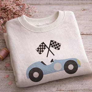 Pode incluir: Camisola branca com um design de carro de corrida azul claro, rodas pretas e duas bandeiras quadriculadas cruzadas. A camisola tem gola redonda. Um design divertido e lúdico para crianças.