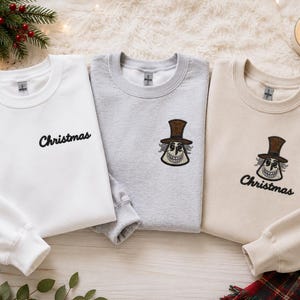 Puede incluir: Tres sudaderas de cuello redondo en blanco, gris claro y beige. La sudadera blanca presenta la palabra Christmas bordada en negro. La gris muestra un personaje bordado con sombrero de copa. La beige incluye ambos elementos bordados.