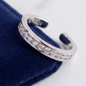 May include: A silver-toned open ring featuring a channel setting with a row of clear, round-cut stones. The inner band is clearly stamped with the text S925, indicating the metal composition. The ring has a polished finish and an open design.