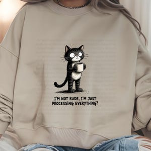 May include: Beige sweatshirt featuring a black and white cartoon cat holding a coffee mug. The cat has large eyes and a slightly grumpy expression. Text on the sweatshirt reads, "I'M NOT RUDE, I'M JUST PROCESSING EVERYTHING?"