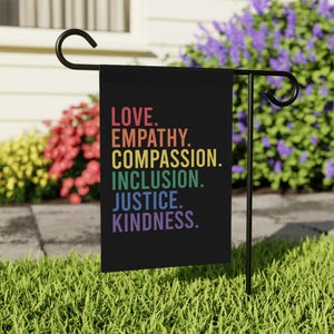May include: A black garden flag with the words "LOVE. EMPATHY. COMPASSION. INCLUSION. JUSTICE. KINDNESS." in rainbow colors. The flag is displayed on a black metal stand in a garden setting with colorful flowers.