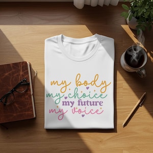 Puede incluir: Camiseta blanca con el texto "my body my choice my future my voice" en escritura naranja, verde, morada y rosa. La camiseta está doblada sobre una superficie de madera, con un cuaderno, gafas, un bolígrafo y una taza de café.