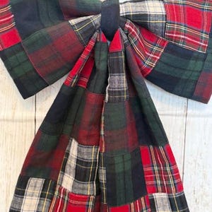 May include: A large, decorative bow made of patchwork fabric in a classic plaid pattern. The bow features a combination of red, green, black, and cream-colored squares, with a dark center and a jute string for hanging. The bow is a festive decoration.