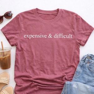 May include: A dusty rose-colored t-shirt with the words "expensive & difficult" printed in white. The shirt is paired with blue jeans, sunglasses, and a drink in a clear plastic cup with a straw.