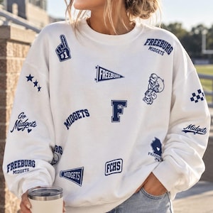 Puede incluir: Una sudadera blanca de cuello redondo con varios estampados gráficos en azul marino que incluyen el texto Freeburg Midgets, FCHS, un dedo de espuma número uno, un banderín, un megáfono, una mascota, estrellas, un patrón de cuadros y un mapa.