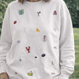 Könnte beinhalten: Cremefarbenes Sweatshirt mit aufgestickten Patches verschiedener Objekte, darunter ein Buch, ein Cocktail, der Buchstabe "A", ein Football, eine Schleife, ein Hund, eine Muschel, ein Hummer, eine Lilie, ein Hockeypuck, ein Golfplatz und eine Brille. Die Buchstaben "STL" sind ebenfalls gestickt.