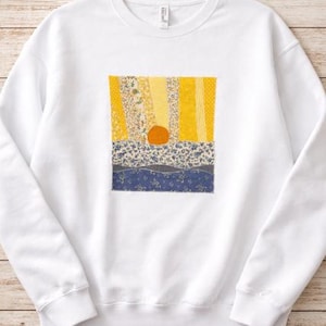 May include: White sweatshirt featuring a colorful patchwork design. The design includes yellow, blue, and floral patterns, with an orange circle in the center. The sweatshirt is made of a soft material and is suitable for casual wear.