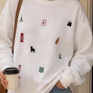 Op de afbeelding: Een wit sweatshirt met ronde hals en kleine geborduurde iconen verspreid over de voorkant, waaronder de Eiffeltoren, een rode telefooncel, een zwarte hond, een boek, een fles, een handtas en een frisdrankblikje, met lange mouwen en boorden.