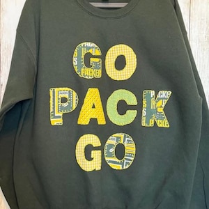 May include: Dark green sweatshirt with the words "GO PACK GO" appliqued on the front. The letters are made of various green and yellow patterned fabrics, including gingham and Packers-themed prints. The sweatshirt has a crew neck and long sleeves.