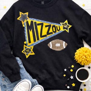 May include: Black sweatshirt with a yellow and blue "MIZZOU" design, stars, and a football graphic. The sweatshirt is paired with blue jeans and white sneakers. A yellow pom-pom and coffee cup are also visible.