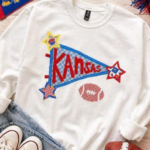 Puede incluir: Sudadera blanca con un diseño de banderín que dice "Kansas" en letras rojas. El banderín es azul con un patrón de cuadros. La sudadera también presenta parches de estrellas y fútbol. Un par de zapatillas blancas y unos vaqueros azules están en la imagen.
