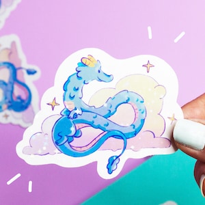 May include: A blue and white sticker featuring a cartoon dragon curled up on a cloud with stars. The dragon has a light blue body with white accents and a yellow horn.