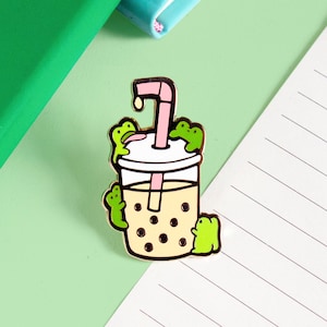 May include: Gold-tone enamel pin featuring a boba tea cup with a pink straw and four green frogs surrounding it.