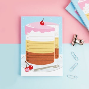 May include: A pink, yellow, and brown layered cake with a cherry on top, on a white plate with a fork. The cake is on a blue and white notepad with a dotted background. The notepad has a white border and a white line for writing.