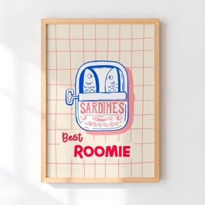 May include: A framed art print featuring a whimsical illustration of a sardine can with two fish inside. The can is blue and pink, with the word "SARDINES" in pink. Below, the words "Best Roomie" are in red.