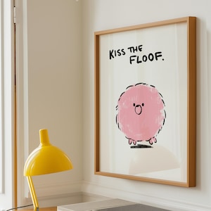 Pode incluir: Impressão artística emoldurada com uma ilustração de uma criatura rosa e fofa. O texto "Kiss the Floof" está em preto. A moldura é de madeira clara, ideal para um quarto infantil.