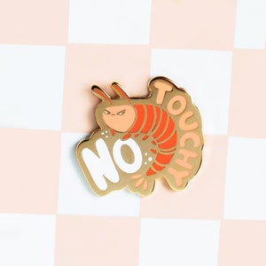 May include: Gold-coloured enamel pin featuring a cartoon shrimp with angry eyes and the words "No Touchy" in a white outline.