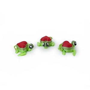 May include: Three green turtle figurines with red heart-shaped shells. Each turtle has black eyes and a small loop for attachment. The turtles are arranged on a white background.
