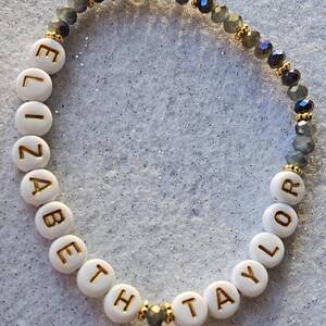 May include: A beaded bracelet featuring white circular beads with gold lettering spelling out ELIZABETH TAYLOR, interspersed with small gold-toned spacers and a section of faceted, dark grey, translucent beads arranged in a circular pattern.
